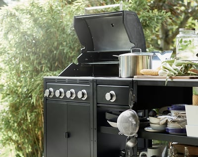 Black gas grill with stainless steel burners & handles, large cooking surface, side shelves, and built-in thermometer, surrounded by greenery.
