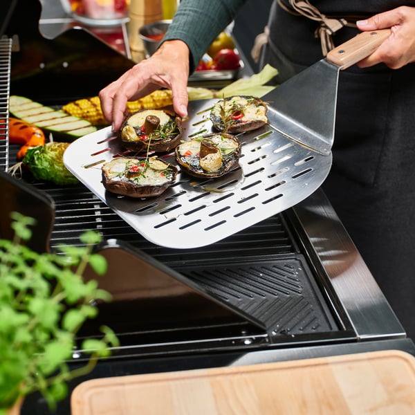 Stainless steel griddle spatula lifting grilled veggies from bbq, showing its non-stick and versatile cooking use.