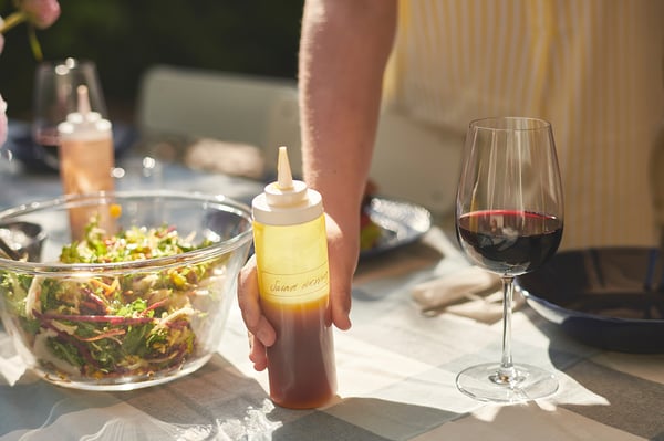 Hand holds dressing bottle labeled jamaican near salad bowl and wine glass on a table.