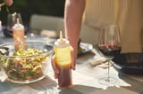 Hand holds dressing bottle labeled jamaican near salad bowl and wine glass on a table.