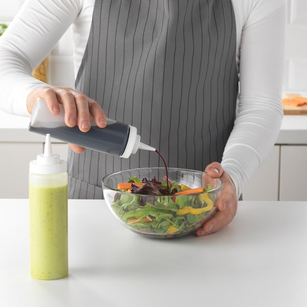 A person pours dressing from a gray squeeze bottle onto a salad in a bowl on a kitchen counter.