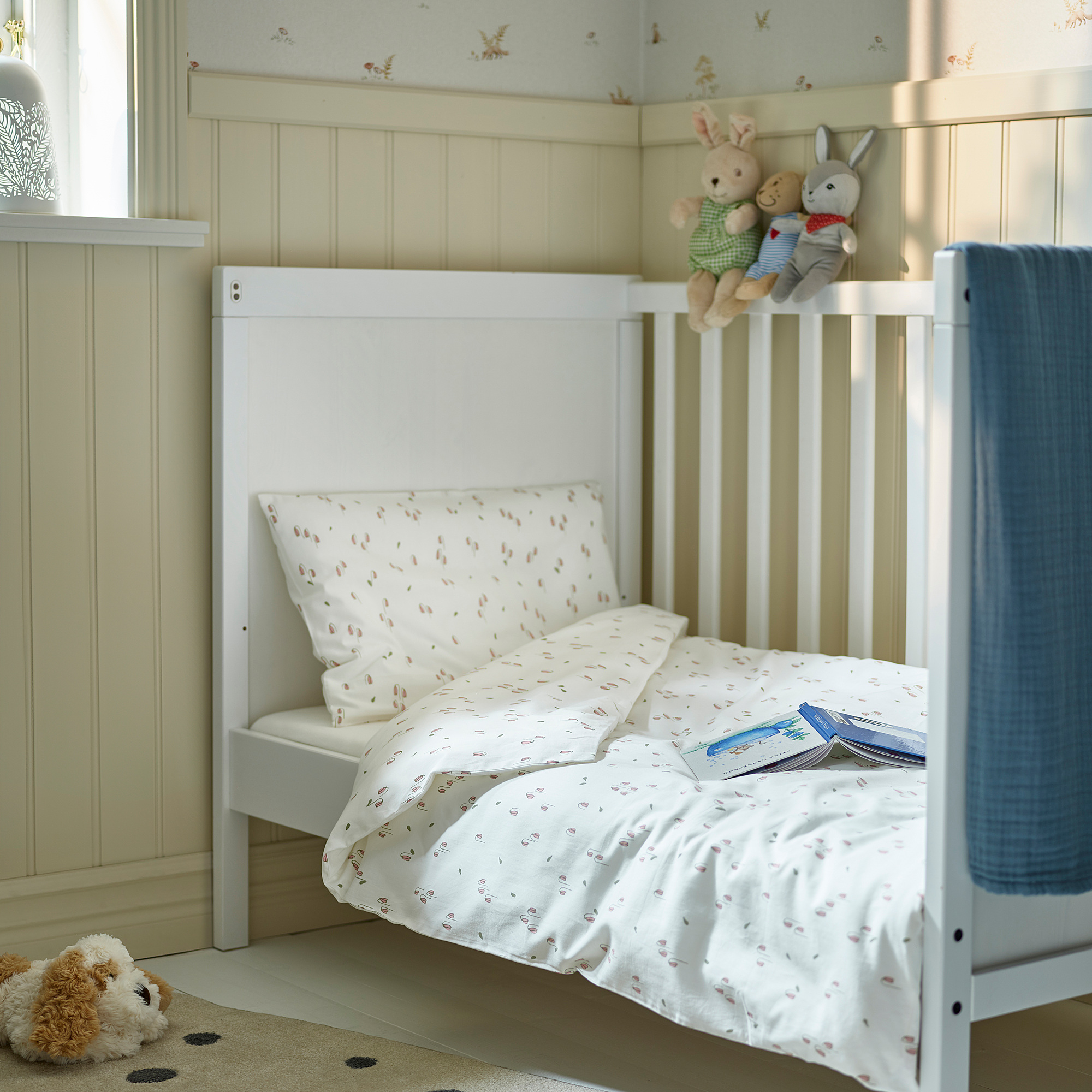 Small white crib with pink flowered linens, plush toys, and a book on top; set against pastel green walls with floral patterns.