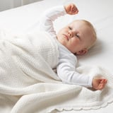 Baby lying on white blanket, arms raised. Soft, knitted cotton cover, suitable for naps.