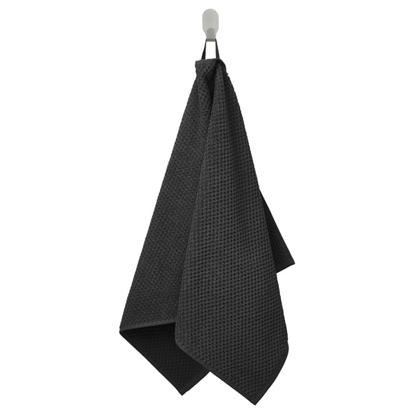 A black GULVIAL towel hangs on a hook. Made of 100% cotton, its soft and fluffy for comfort and absorption.
