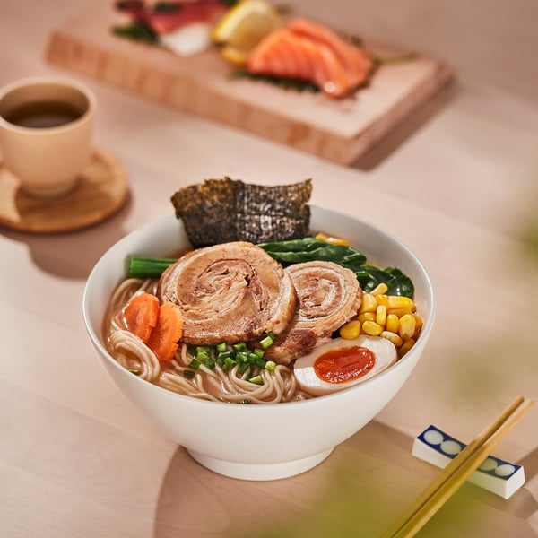 A bowl of ramen noodles with vegetables and meat; chopsticks and tea beside it.