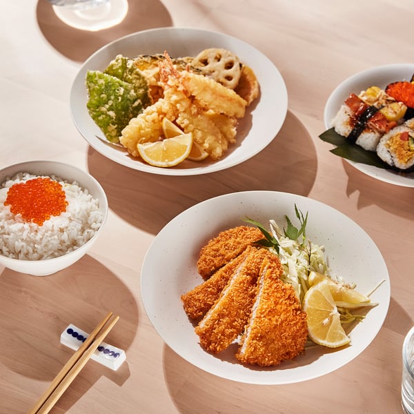 Four dishes of japanese cuisine on light wood table, including tonkatsu, sushi, and tempura, served on stackable porcelain bowls.