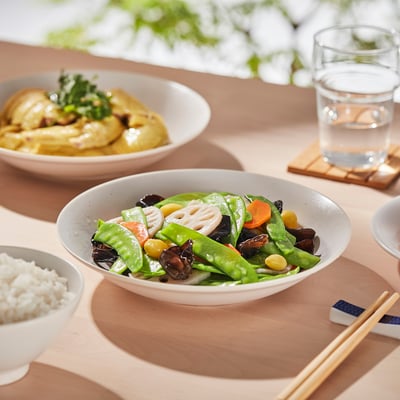 Three bowls of food on a wooden table: stir-fried vegetables, chicken with greens, and rice. Chopsticks and water glass nearby.