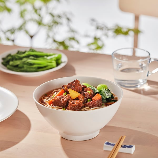 A bowl of beef noodle soup with vegetables on a wooden table, accompanied by a side dish of greens, a glass of water, and chopsticks.