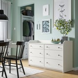Modern bedroom with white HAUGA chest, black chairs, light blue walls, nature prints, plant, and closet.