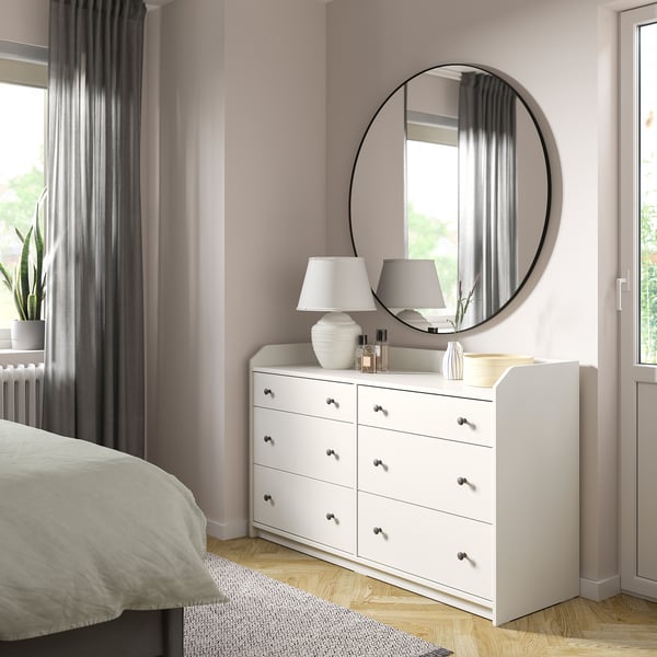 Depicts a white HAUGA chest of drawers with six drawers in a bedroom setting. Atop is a circular mirror, white lamp, and decorative items.