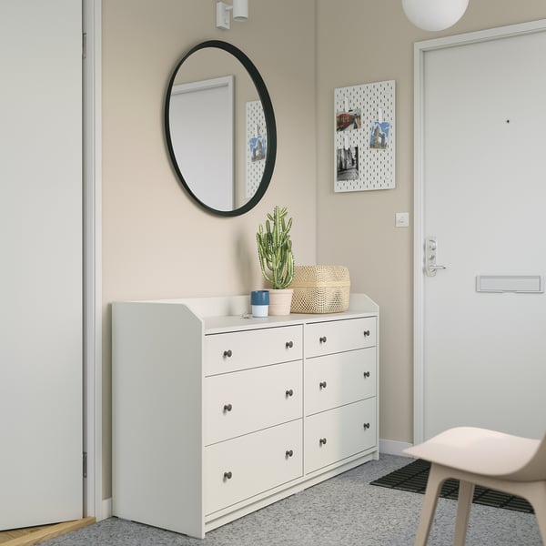 Neutral-colored häuga dresser by entrance, six drawers, round mirror above, plant, and pegboard on wall.