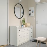 Neutral-colored häuga dresser by entrance, six drawers, round mirror above, plant, and pegboard on wall.
