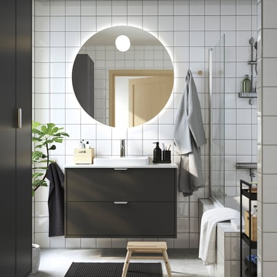 Modern bathroom: HAVBÄCK black unit, round mirror, wood stool, towels, plant, glass shower.