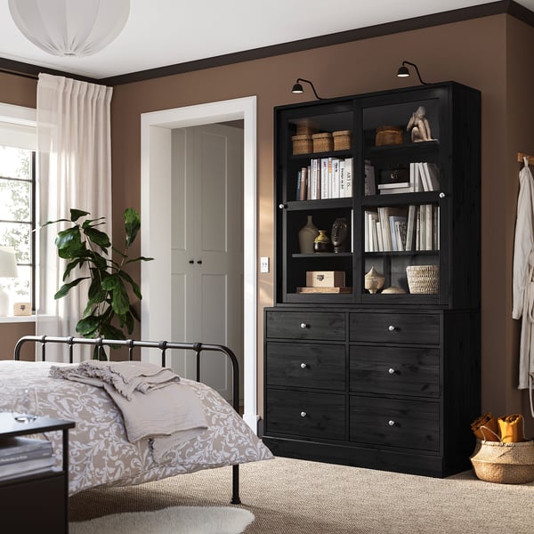 Modern bedroom with dark wood dresser, glass bookcase, metal bed, soft lighting, and plant. Beige and brown tones.