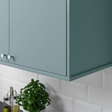 Blue kitchen cabinet with round knobs, hiding lighting unit, placed above white tile backsplash with small green plant.