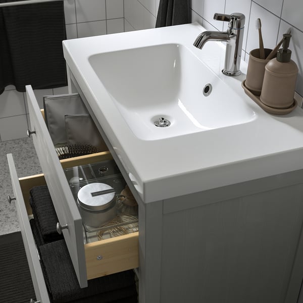 A bathroom vanity with a white sink and gray cabinet. The drawer is open, showing storage solutions like compartments and a bin.