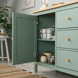 Green HEMNES sideboard with open door displaying cups, plates, and jars on shelves, showing storage and organizational.
