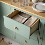 Open green sideboard showing neatly folded napkins in drawer, with wooden handles and countertop displaying glasses and plates.