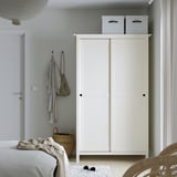 HEMNES wardrobe: tall, white, sliding doors, tops with 2 gray boxes. Next to bed, above basket, chair.