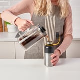 Blonde woman pouring coffee from a clear glass jug into a silver and black insulated thermos.