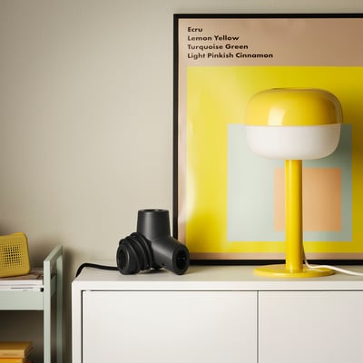 Black HORNFELS 4-way extension cord sits on white furniture, near a yellow lamp. Small, compact, creative design.