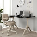 Black IDÅSEN desk, white legs, beige chair. Laptop, mug, lamp, pen holder, clock, wall photos.