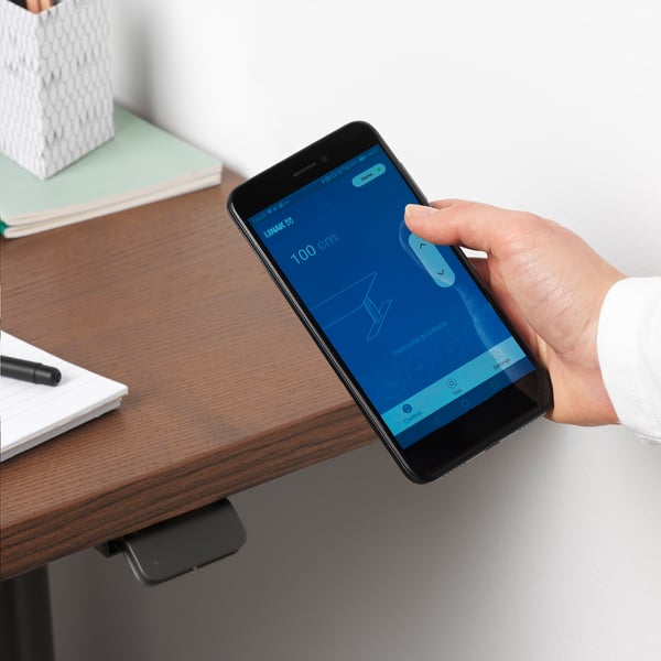 Hand holds smartphone adjusting height of IDÅSEN desk, app visible. Desk varies from 63-127 cm, with cable management underneath.