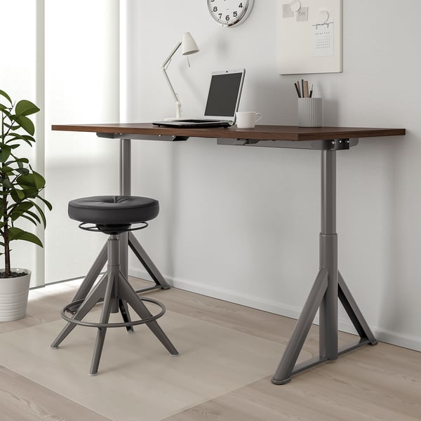 IDÅSEN sit-stand desk with laptop, lamp, and cup. Black chair beneath, light flooring, white walls, side plant.