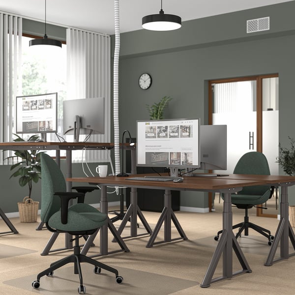 Modern office with dual wooden desks, multiple monitors, green chairs, and efficient layout against a green wall with windows.