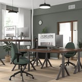 Modern office with dual wooden desks, multiple monitors, green chairs, and efficient layout against a green wall with windows.