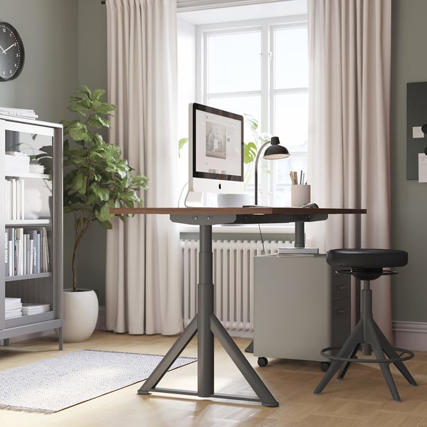 Black adjustable desk in home office with stool, monitor, lamp, plants, bookshelf, and window.