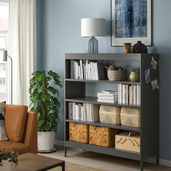 Black IDÅSEN bookshelf with adjustable shelves against blue wall, holding books and decor, near orange chair and plant.
