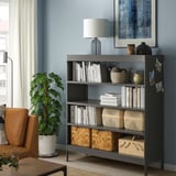 Black IDÅSEN bookshelf with adjustable shelves against blue wall, holding books and decor, near orange chair and plant.