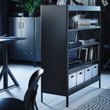 Modern black office setup featuring IDÅSEN metal shelves with storage boxes, a desk, and a plant.