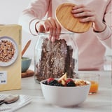 Placing bamboo lid on jar of bread. Table has fruit, oj, ikea box, spoon.
