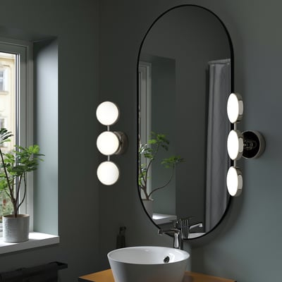 Large black oval mirror with modern lights on either side. The sleek design contrasts against a dark wall. A small sink is nearby.