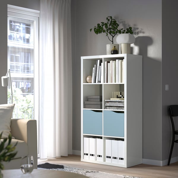 White KALLAX bookshelf with blue door, near window, holds books, decor, plants, larger than chair.