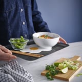 Person holding chopsticks near tray with bowl, edamame, and cutting board with lime and herbs.