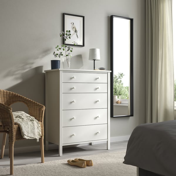 Minimalist bedroom with white KILBERGET tall chest, lamp, vase, wicker chair, and large mirror.