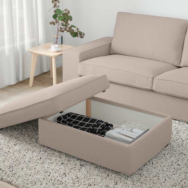 Beige KIVIK footstool with storage, magazines, blanket, next to sofa and side table with vase and teacup. Bright, airy window.