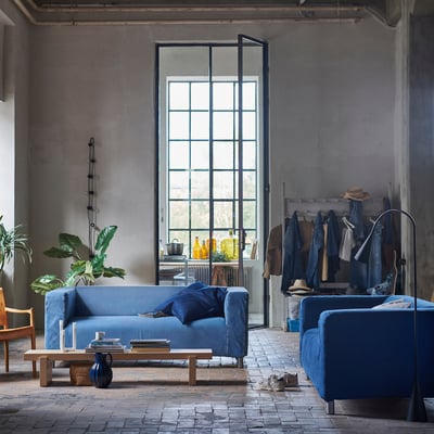 Modern industrial living room with blue KLIPPAN sofa & armchair, wooden coffee table, large window, and plant.