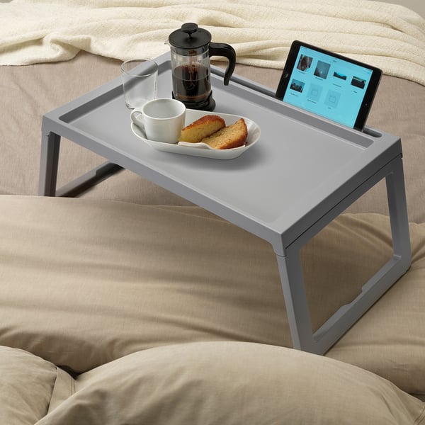 A gray bed tray holds a coffee maker, cup, food, and tablet on beige bed linens.