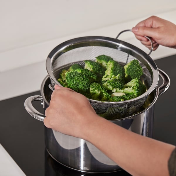 Two hands using a KLOCKREN colander to drain bright green broccoli over a pot, demonstrating its straining function.