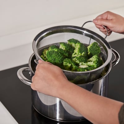 Two hands using a KLOCKREN colander to drain bright green broccoli over a pot, demonstrating its straining function.