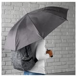 Person holding black KNALLA umbrella, white sleeves visible, against grey brick wall.