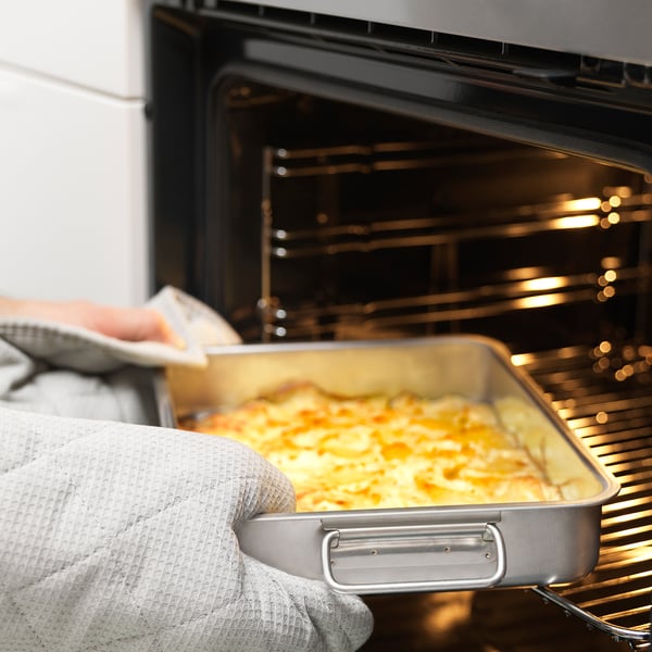 A person wearing oven gloves removes a roasting tin filled with food from an oven. The tin has handles and is suitable for both oven and grill use.