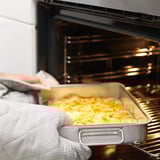 A person wearing oven gloves removes a roasting tin filled with food from an oven. The tin has handles and is suitable for both oven and grill use.