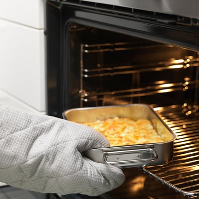 Hand holding metal dish with cooked food, placing it into oven, demonstrating oven-safe feature.