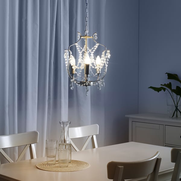 Dining chandelier with crystals and white chairs, bulbs glowing, vase, glasses on raffia, sheer curtains.