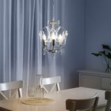 Dining chandelier with crystals and white chairs, bulbs glowing, vase, glasses on raffia, sheer curtains.